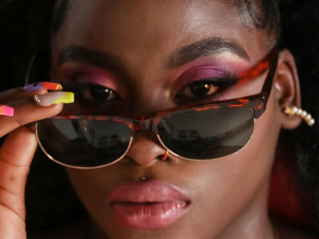 NaomiLoveHot with dark skin, pink and purple eyeshadow, brown tortoiseshell sunglasses, neon yellow and pink nails, gold hoop earrings, and a nose piercing, looking serious.