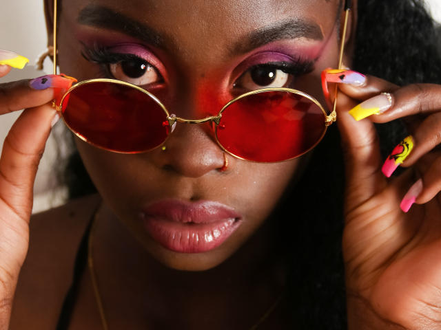 NaomiLoveHot with dark skin, pink and purple eyeshadow, red-tinted sunglasses, yellow and pink nails, holding glasses, shiny pink lips, and long black hair.