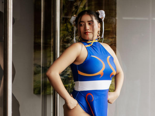 Veronica, les cheveux noirs attachés en chignons, portant une robe bleue et jaune, les mains sur les hanches, debout devant une porte en verre, montrant ses seins et une petite fesse..