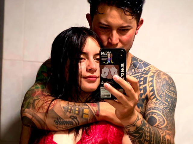 TravesiasSexuales, met nat zwart haar, een rode kantbeha en tatoeages, houdt een telefoon vast terwijl een getatoeëerde man met donker haar haar nek kust. Zijn armen zijn om haar heen..