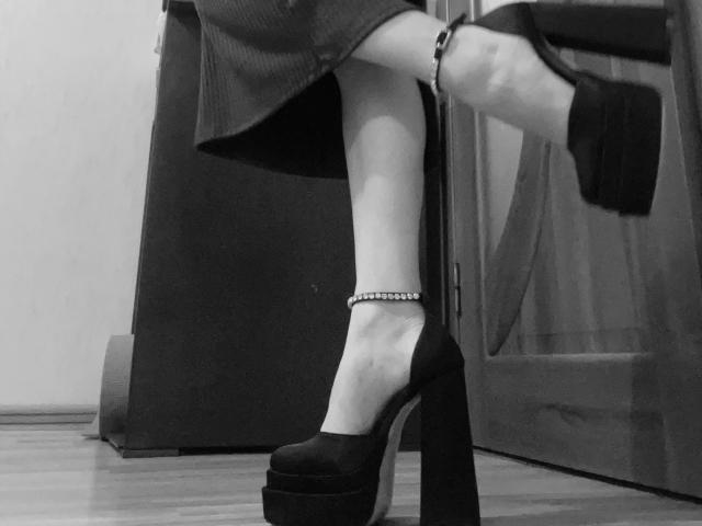 SugarrX's foot in a black platform heel with a studded ankle bracelet, leg in a dark skirt, standing on a wooden floor, door in the background.