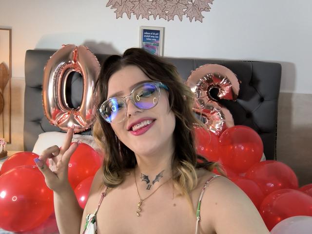 KimSummer with pink-tinted glasses, tongue out, holding rose gold "20" balloons, surrounded by red balloons, wearing a floral top, showing a black tattoo on her neck.