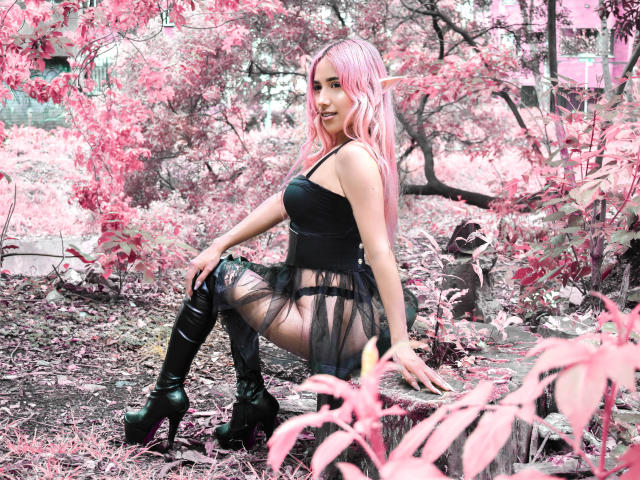 BrianaHarpers with pink hair, elf ears, black corset, sheer black dress, black thigh-high boots, squatting on log, surrounded by pink trees, showing her ass and pouty face.