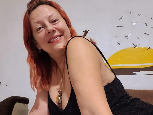 BarbaraBowie with red hair, smiling, wearing a black sparkly dress, showing her tits, necklace with shell and bead, sitting on brown couch, birds in wall art.