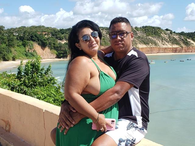 CaboLara with big ass in green dress, arms around CaboLara with black shirt, white shorts, holding pink phone, standing by blue water, green hills, cloudy sky.
