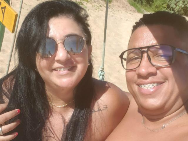 CaboLara met zwart haar, een zonnebril in aviatorstijl en een tatoeage, en CaboLara met een bril, die glimlachend naar de camera kijkt, beiden zonder bovenkleding op een zonnig strand..