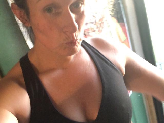 RDreamzCumTru, puffy-lipped, in a black tank top, showing her titts and muscular arms, with her head tilted slightly and brown hair pulled back. Light casts a shadow on her.