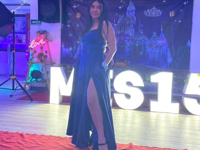 EliMartinez in a blue, high-slit dress, standing on a red carpet, with hands in pockets, black heels, long dark hair, and a confident smile, in a brightly lit room.
