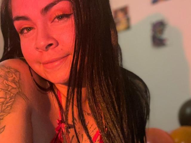 EliMartinez with long black hair, red eyeliner, and a tattoo on her left arm, wearing a red halter top, smiling slightly with red lighting, and a nose piercing.