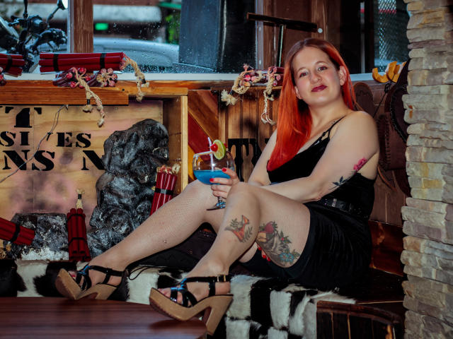 VioletaMora with red hair, black dress, tattoos on thigh and arm, holding blue cocktail, black heels, sitting on wooden bench, stone wall on right, window behind.