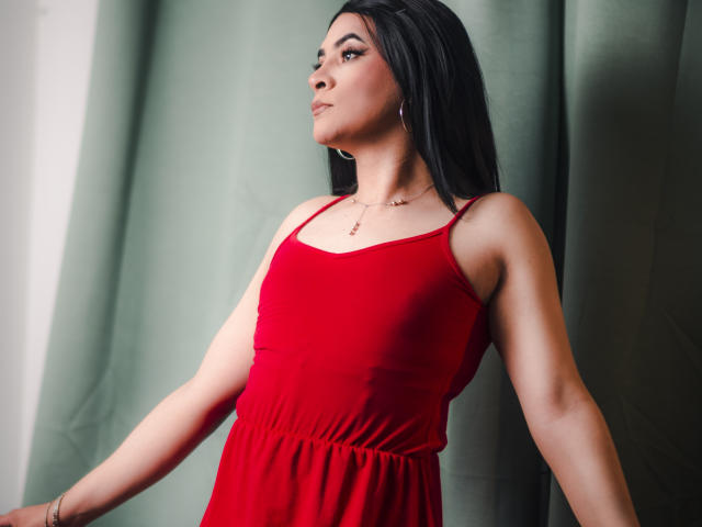 Sara Winson, avec ses longs cheveux noirs, sa robe rouge et ses boucles d'oreilles, regarde sur le côté avec un sourire discret, debout devant un rideau vert. Elle a une belle silhouette..