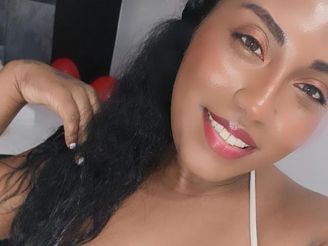 ChocolateTits met donker haar, bruine ogen en roze lippenstift, lachend naar de camera, haar vastgehouden met één hand, een witte top met bandjes aan, haar schattige gezicht laten zien..