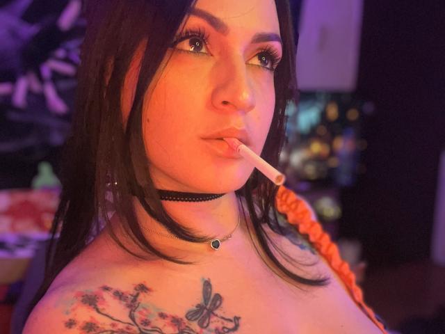 ElinaJhonson with dark hair, green eyes, and a cigarette in her mouth, wearing a black choker, has a butterfly tattoo on her shoulder, and orange braided hair.