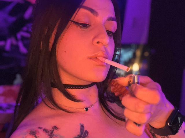 ElinaJhonson with long brown hair, wearing a black choker, smoking a cigarette, tattoos on her chest, pink and purple lighting, holding a silver ring, and a watch on her wrist.