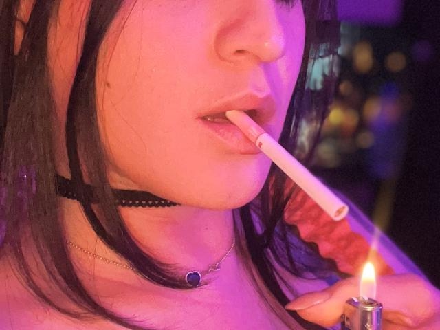 ElinaJhonson with black hair, wearing a black choker, has a lit cigarette in her pink-lit mouth, holding a small candle in her right hand.