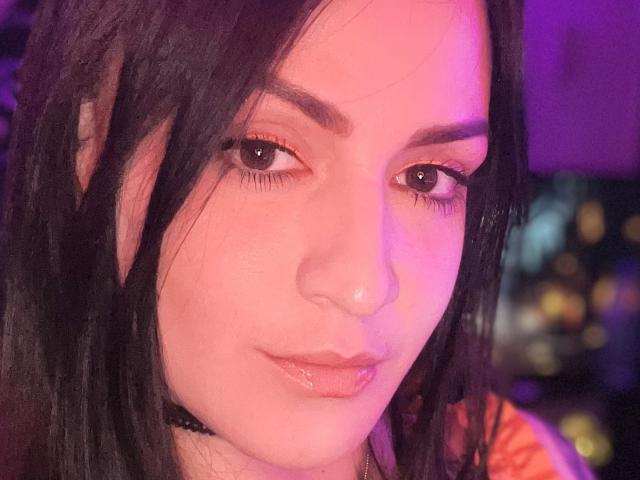ElinaJhonson with pinkish-purple lighting, glossy pink lips, black choker, long black hair, brown eyes, subtle makeup, and a slight smirk. Her nose is small and her skin is fair.