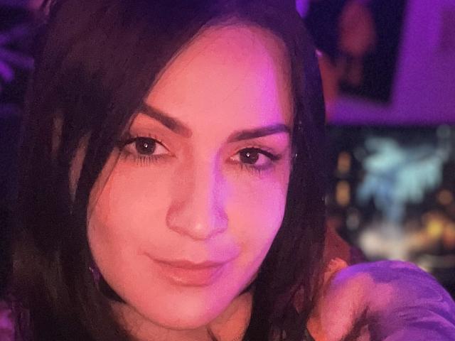 ElinaJhonson with pink and purple lighting on her face, dark wavy hair, and a subtle smile. She's wearing a nose ring and has her hand near her chin.