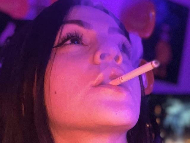 ElinaJhonson with a cigarette in her mouth, purple-lit face, and dark hair, looking up with her lips slightly parted, showing a small mole on her right cheek.