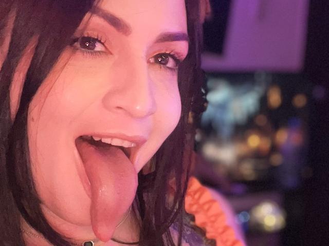 ElinaJhonson's face with pinkish skin, dark brown eyes, and thick eyebrows, tongue fully out, looking goofy. She has dark brown hair, and a small beauty mark on her left cheek.