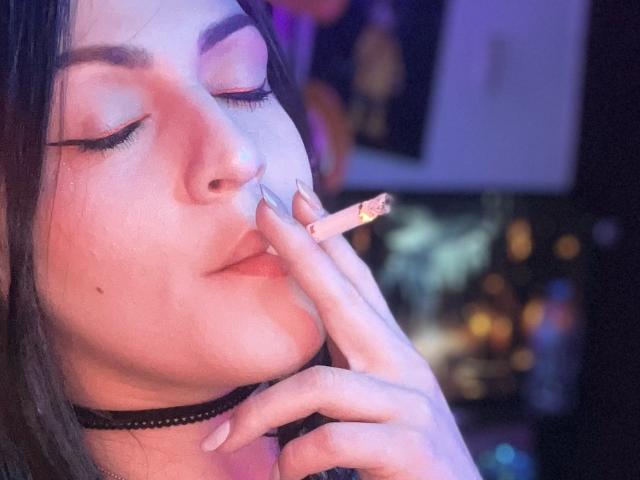 ElinaJhonson with pink eyelids, holding a lit joint to her pink lips, wearing a black choker, her finger with long nails touching her lips. Blurred city lights in background.