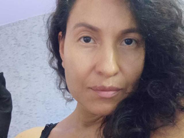 EroticSasha with curly black hair, medium brown skin, and pouty pink lips, wearing a black lace top, looking straight at the camera with a slight, confident smile.