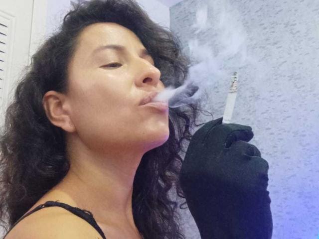 EroticSasha, con cabello negro rizado, labios carnosos y guantes negros, fumando un cigarrillo, con humo blanco saliendo de su boca, vistiendo una blusa negra de encaje..