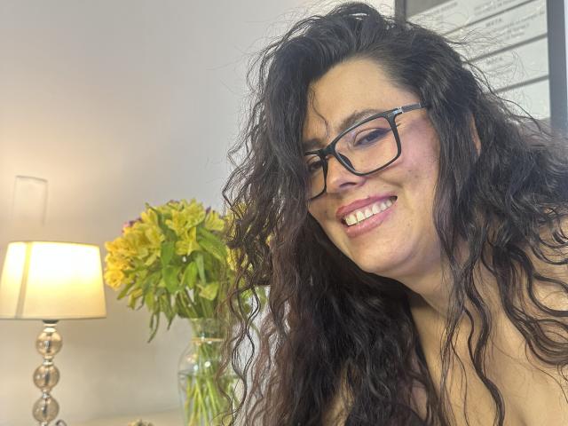 Marthina Baker, con i capelli neri ricci, che indossa gli occhiali, sorride, mostrando i denti, ha la pelle chiara ed è senza top, con un bouquet di fiori gialli e una lampada sullo sfondo..