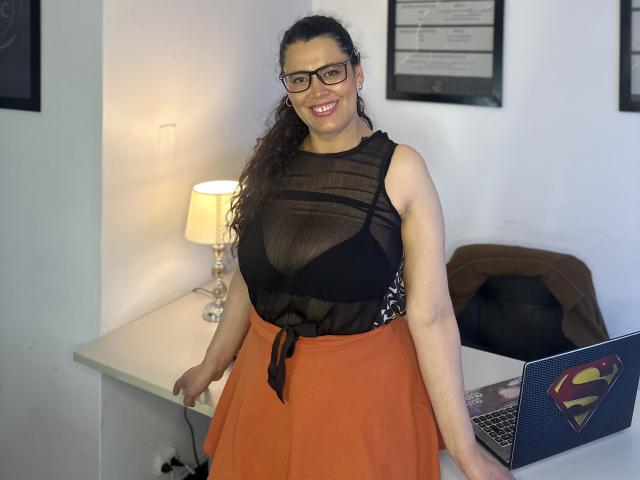 Marthina Baker, con cabello castaño rizado, gafas, una blusa negra transparente que muestra sus senos, una falda naranja, sonriendo, de pie junto a un escritorio con una computadora portátil y una lámpara..