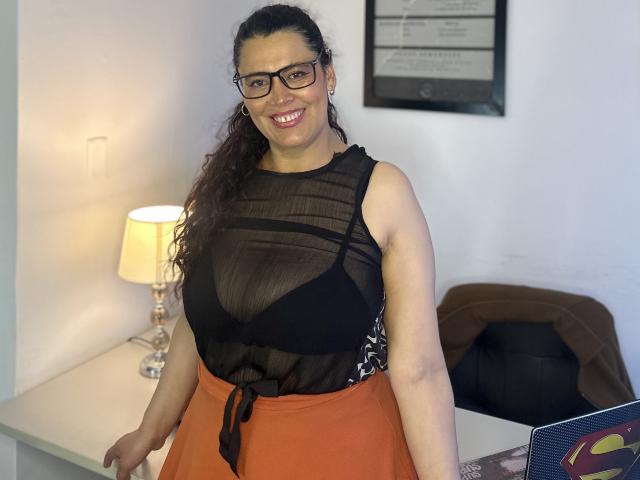 Marthina Baker, com cabelos castanhos cacheados, óculos e um sorriso, vestindo uma blusa preta transparente que mostra seus seios e uma saia laranja, está em pé ao lado de um abajur..