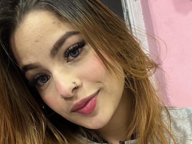 ShantalSummer with blue eyes, nose piercing, and pink lipstick, smiling at the camera, showing off her tits with her long brown hair and light skin.