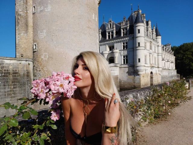 Dominatrix Ana mit langen blonden Haaren, die rosafarbene Blumen hält, trägt ein schwarzes Top mit Trägern, eine goldene Halskette und einen Ring und steht vor einem Schloss, der Himmel ist klar blau..