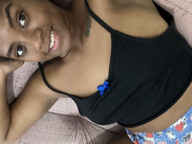 TiffanySammy, avec une peau brun foncé, souriante, portant un débardeur noir avec un nœud bleu et un pantalon de pyjama coloré, allongée sur une surface rose texturée..