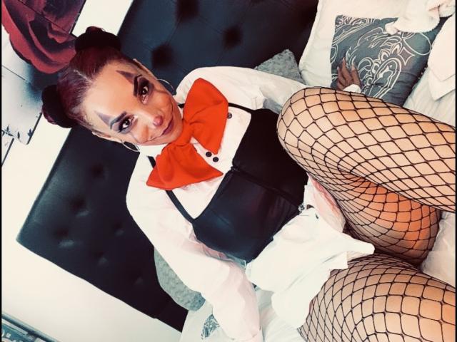 AmberDemons, wearing fishnet stockings, black corset, white shirt, and red bowtie, sits on a bed, legs spread, showing her ass, with a seductive smile.