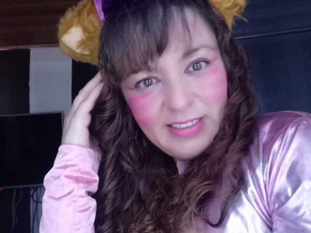 AmandaHotL with pink face paint, brown curly hair, and purple cat ears, wearing a shiny pink top, smiling with her hand in her hair.