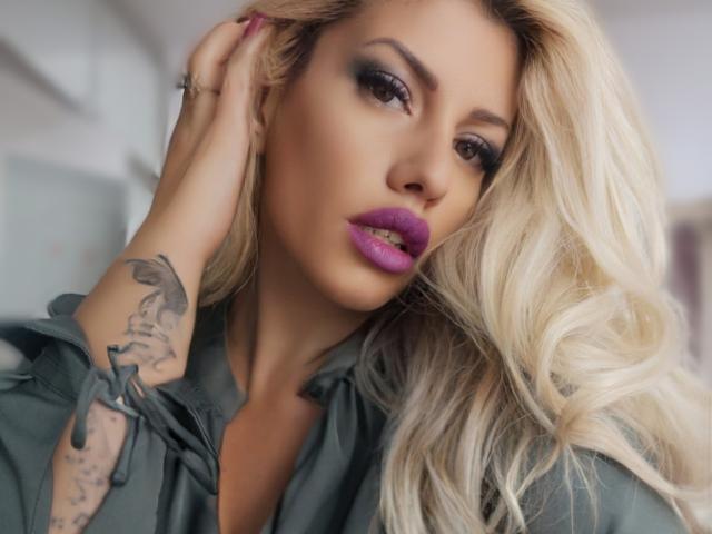 DominatrixAna, com cabelo loiro, batom roxo e uma tatuagem na mão direita, vestindo uma camisa verde, tocando o cabelo com a mão na cabeça..