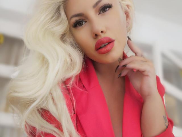 DominatrixAna, con cabello rubio platino, labios rojos y un blazer rosa, luciendo seductora, con la mano cerca de su rostro, uñas largas y un tatuaje en la muñeca..