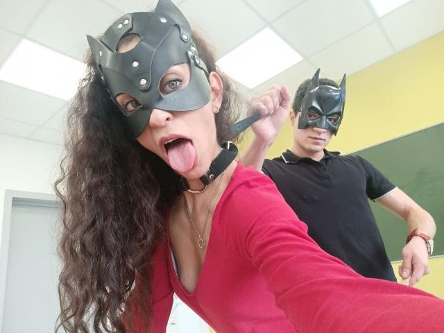 LadyIceJoy avec de longs cheveux bruns bouclés, un haut rouge, un masque noir, la langue tirée, un col noir ; un homme en polo noir, un masque noir, tenant le lanière du masque, derrière elle..