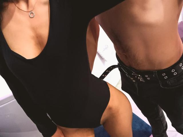LadyIceJoy, vêtue d'une combinaison noire moulante à manches longues avec un décolleté en V plongeant, qui met en valeur ses seins et ses fesses, se tient à côté d'un homme noir, torse nu et portant une ceinture..