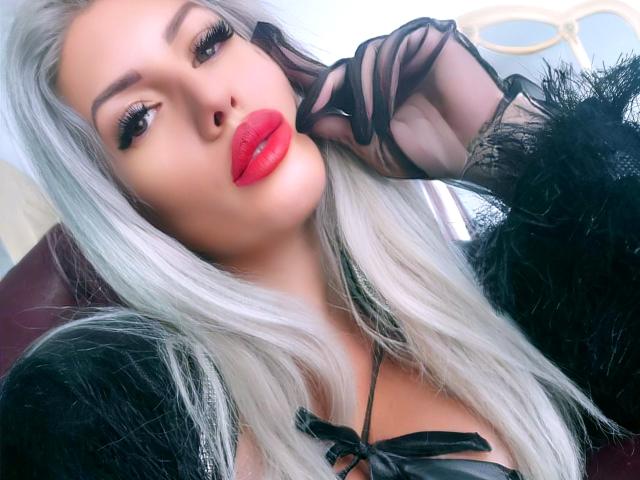 DominatrixAna com cabelo branco comprido, batom vermelho, luvas de malha preta e pele sintética preta, fazendo beicinho com a mão perto do rosto, mostrando os seios em uma blusa de cetim preta..
