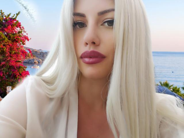 DominatrixAna met platinablond haar, volle roze lippen en een wit shirt, staart in de camera, in de buurt van een felrode bougainvillea en een blauwe oceaan op de achtergrond..