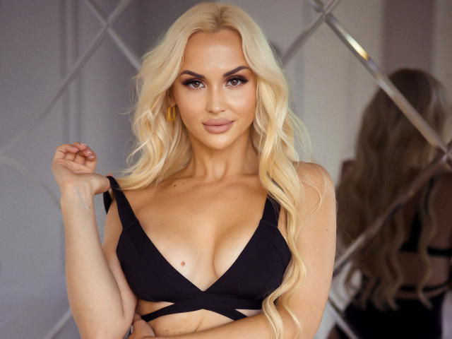 Blonde Klubnika, com cabelo ondulado, vestindo uma blusa preta de decote em V que mostra seus seios, brincos de ouro e um leve sorriso, está em frente a um espelho. Seus olhos estão fixos..