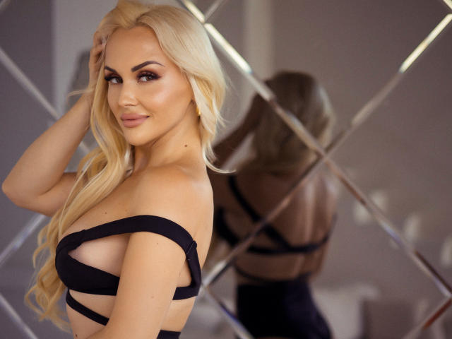 Blonde Klubnika con cabello largo, top negro sin tirantes que muestra sus pechos, sonriendo al reflejo en el espejo, con una mano en el cabello, maquillaje suave, luz cálida, sexy, segura de sí misma, reflejo en un espejo con diseño de diamantes..