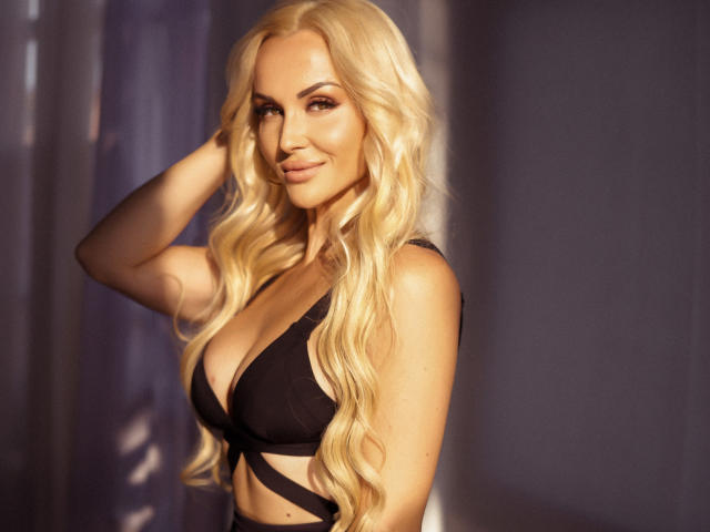 Blonde Klubnika with wavy hair, black bra, and cleavage, smiling, hand in hair, sunlight highlighting her. Boobs are visible, she's sexy and attractive.