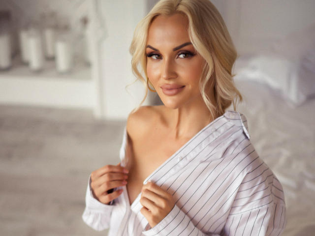 Blonde Klubnika with wavy hair, wearing a white pinstripe shirt, off-shoulder, exposing her right breast, smiling, holding shirt, soft bedroom light, smooth skin, sexy.