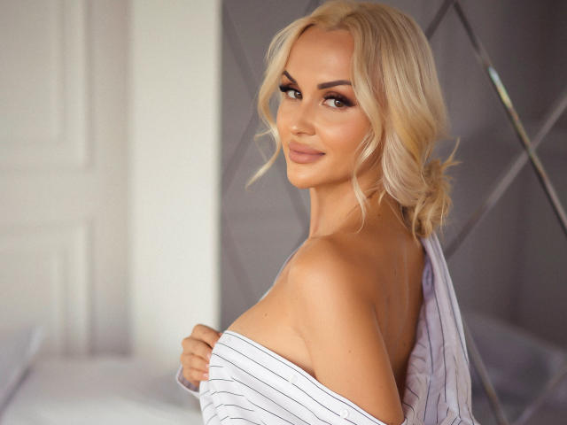 Klubnika with blonde hair, looking over shoulder, smiling, holding striped shirt off her tits, showing her bare back and shoulder. Soft lighting, slightly wavy hair.