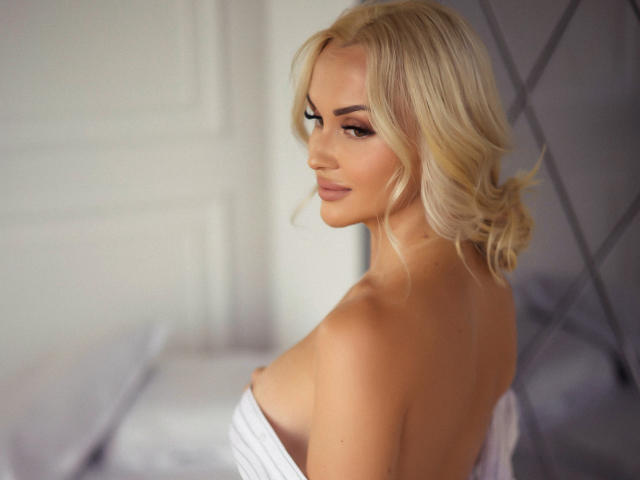 Blonde Klubnika with wavy hair, looking over her shoulder, smiling, topless, exposing her tits, with a white bed and grey diamond-patterned wall in the background.