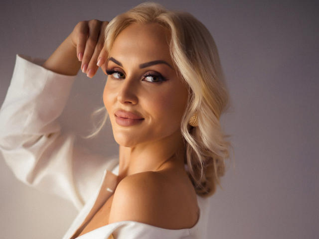 Blonde Klubnika with wavy hair, wearing a white off-shoulder top, smiling at the camera, hand on her head, golden earrings, soft light, looking sexy and confident.