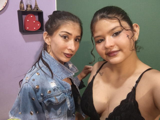 AngelaAndBarbara with dark hair, wearing a denim jacket and black lace bra, showing tits, small nose star tattoo, and brown lipstick, standing side-by-side, smiling.