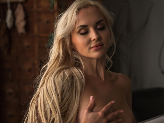 Blonde Klubnika, con i capelli lunghi, gli occhi chiusi e le unghie rosa, copre i suoi seni con una mano, e ha un'espressione sexy e serena. L'illuminazione calda mette in risalto la sua pelle liscia..