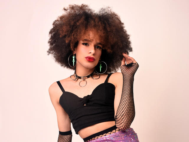 KataleyaFowler with afro, red lipstick, black fishnet gloves, choker, black bra, pink glitter skirt, green earrings, holding afro with right hand, looking at camera.